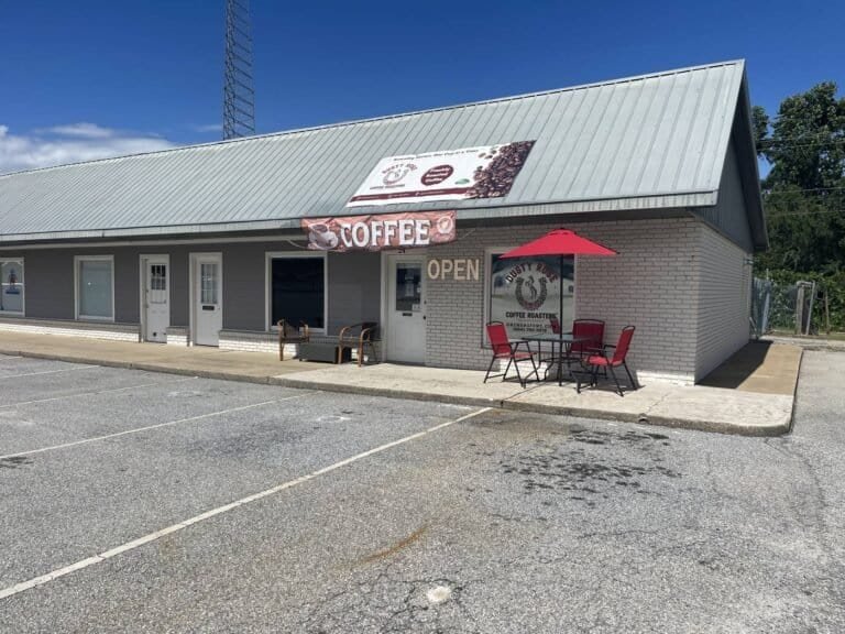 Exterior of a small coffee shop with a metal roof, OPEN sign, and red outdoor seating under a red umbrella.